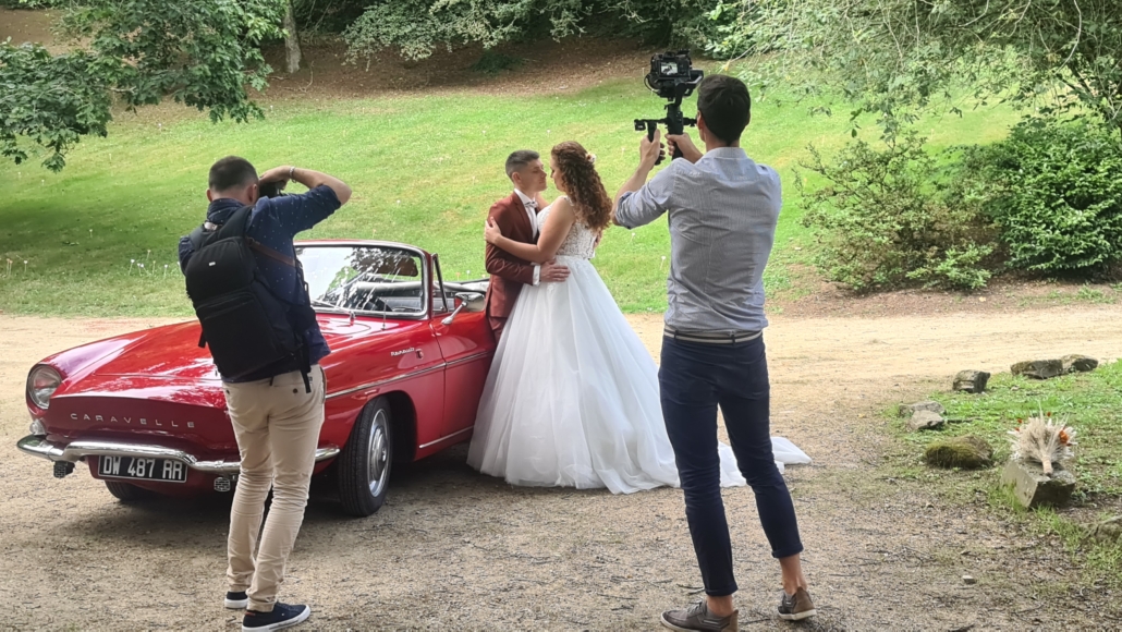 Mariés enlacés contre une décapotable rouge posant pour leurs photographe et vidéaste