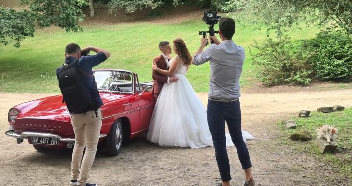 Mariés enlacés contre une décapotable rouge posant pour leurs photographe et vidéaste