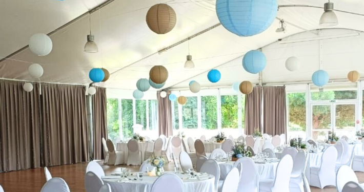 salle de reception de mariage thème bretagne aves tables rondes nappées en blanc, chaises houssée en blanc et lanterne rondes dans des dégradé de bleu et taupe suspendues au plafond