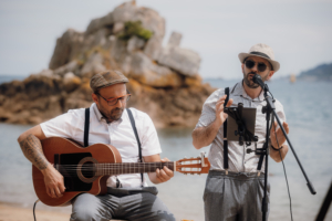 Chanteur et guitariste en cours de prestation sur une plage, la mer et un gros rocher derrière eux lors d'un évènement by Glad events