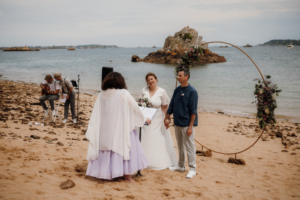 Gwladys, l'officiante de cérémonie laïque de Glad events sur la plage en train d'unir de jeunes époux accompagnés par des musiciens en chanteurs, la mer derrière eux;