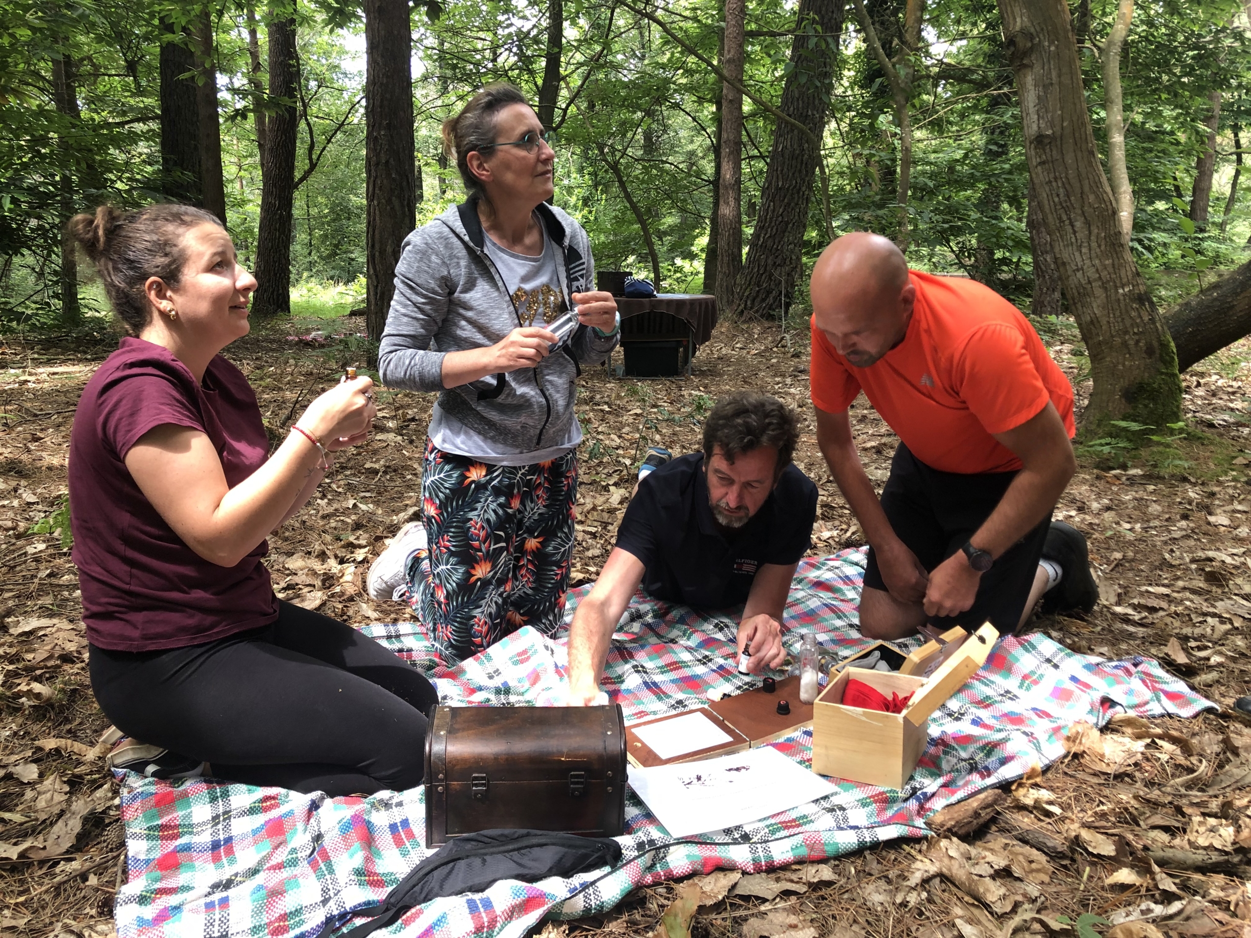 groupe de personnes assises ou à genoux sur un plaid posé par terre dans la forêt. Les deux hommes sont concentrés sur l'énigmes et les deux femme parlent à une personne en dehors du champ de la photo