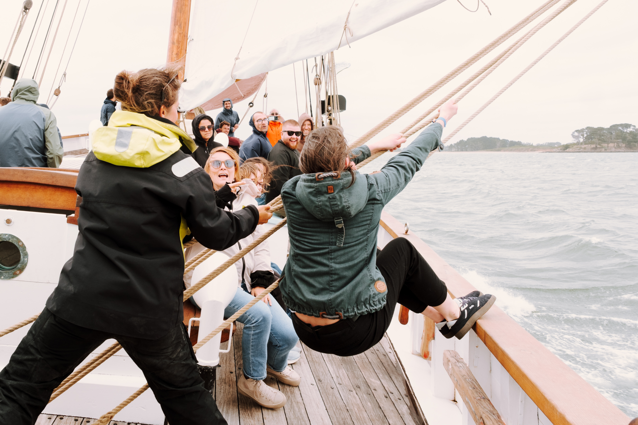 femme à bord d'un bateau lors d'un team building Glad events, tirant sur un bout de totes ses force, les pieds en appui sur la balustre, son équipe en arrière plan l'encourageant et son animateur la guidant à côté