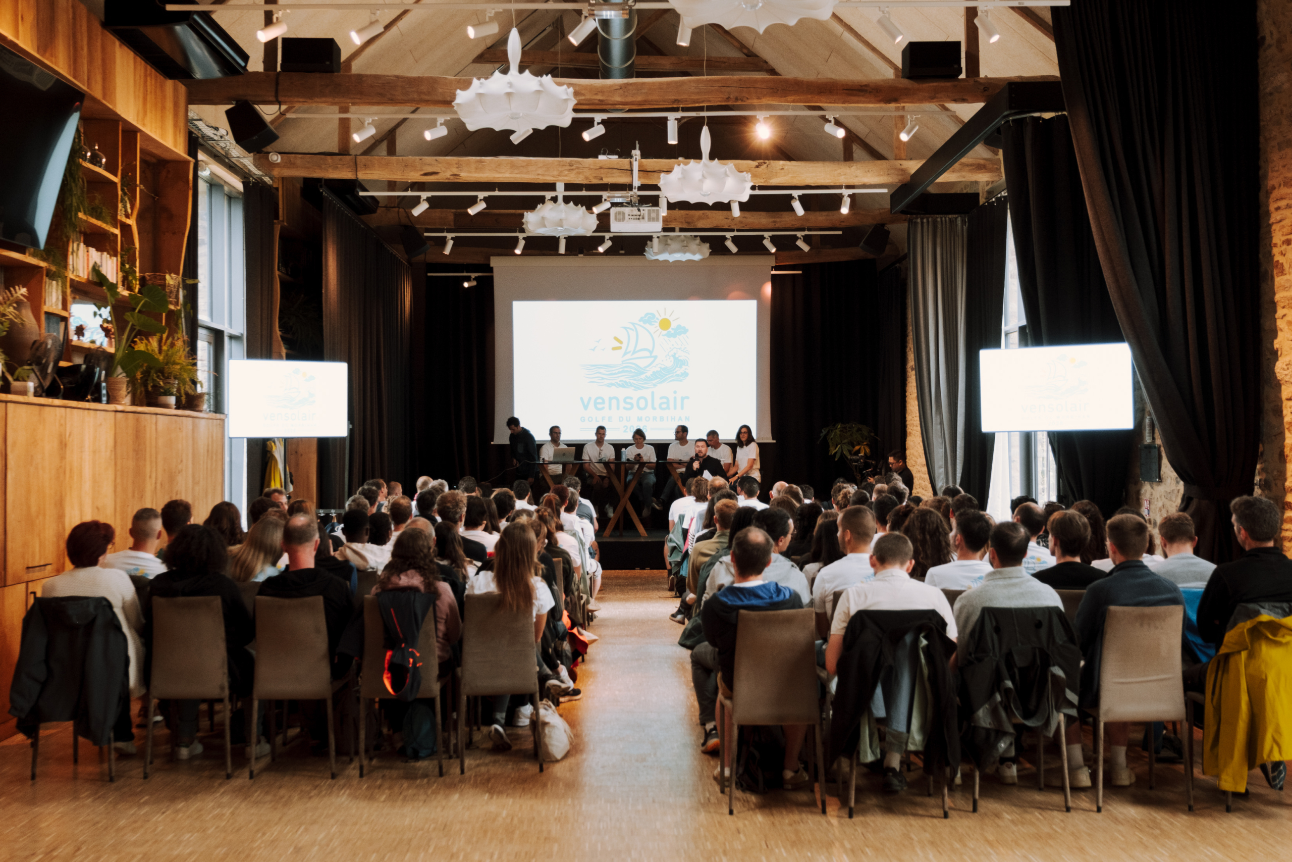 Séminaire résidentiel par Glad events dans une salle haute de plafond, les gens sont photographiés de dos, une allée centrale et un écran blanc sur fond noir, elle est très haute de plafond